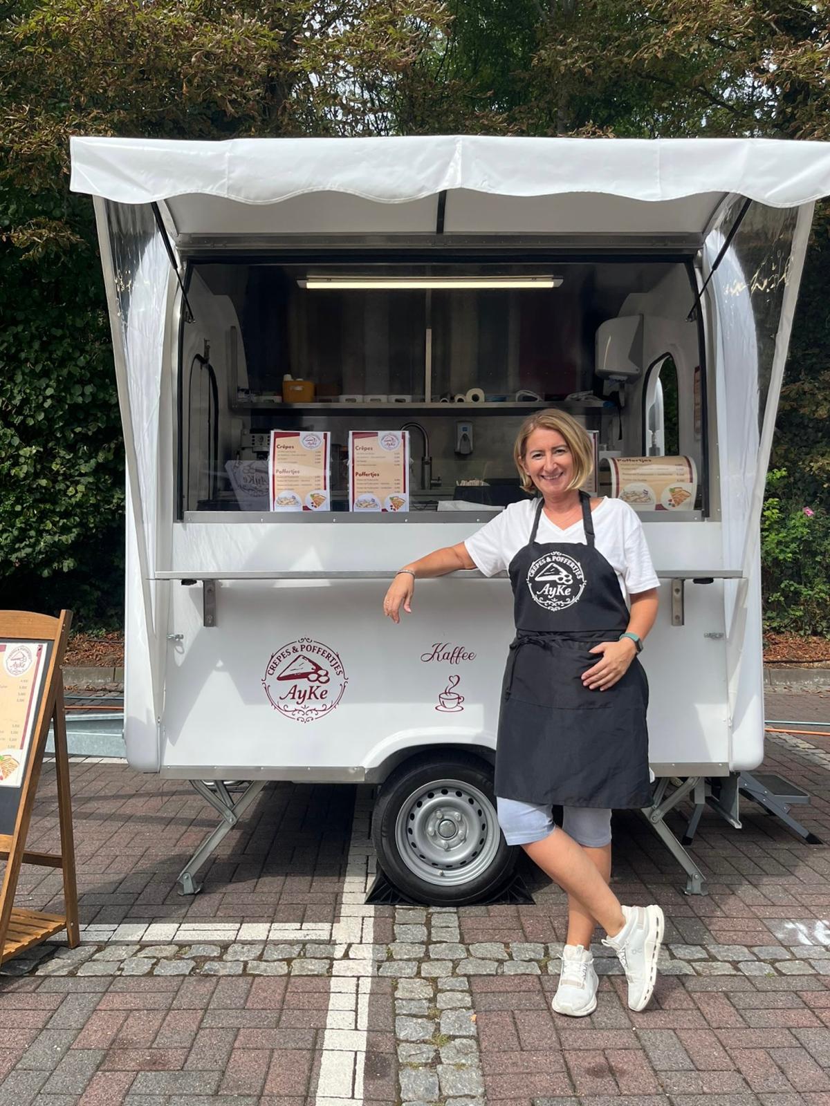 Ay.ke-Foodtruck-Crepes Ay-ke steht in schwarzer Schürze vor ihrem 2 Meter Crepes und Poffertjes Foodtruck in weiß